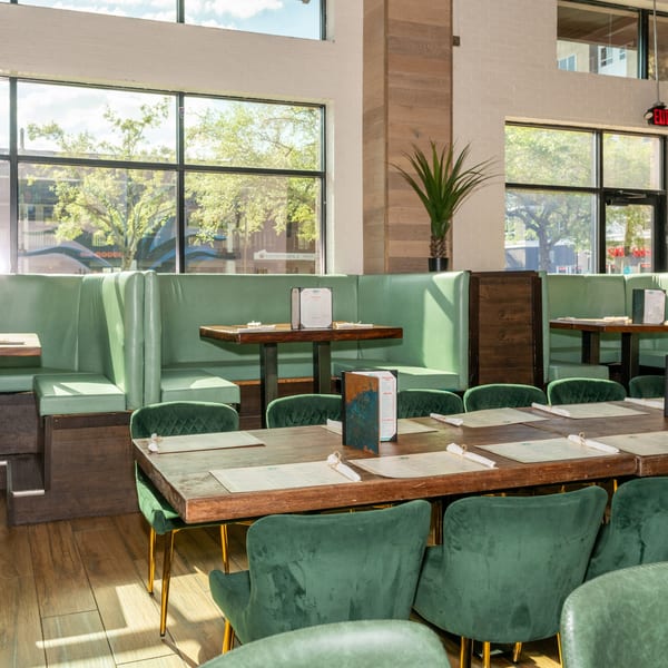 Restaurant interior with green booths, dark wooden tables, and green velvet chairs near large windows.