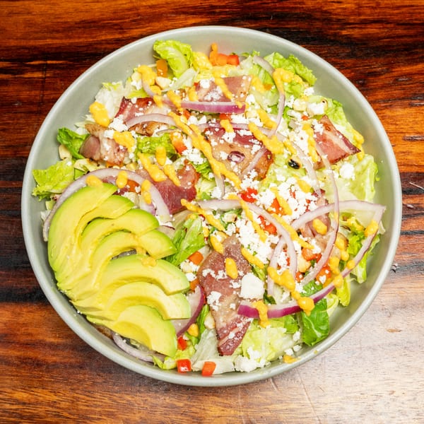 A vibrant salad with lettuce, bacon, red onion, avocado, feta cheese, and a yellow dressing.
