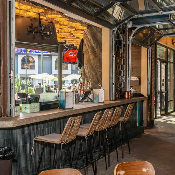 An outdoor bar with wicker stools, bottles, and menus, overlooking a sunny street scene.