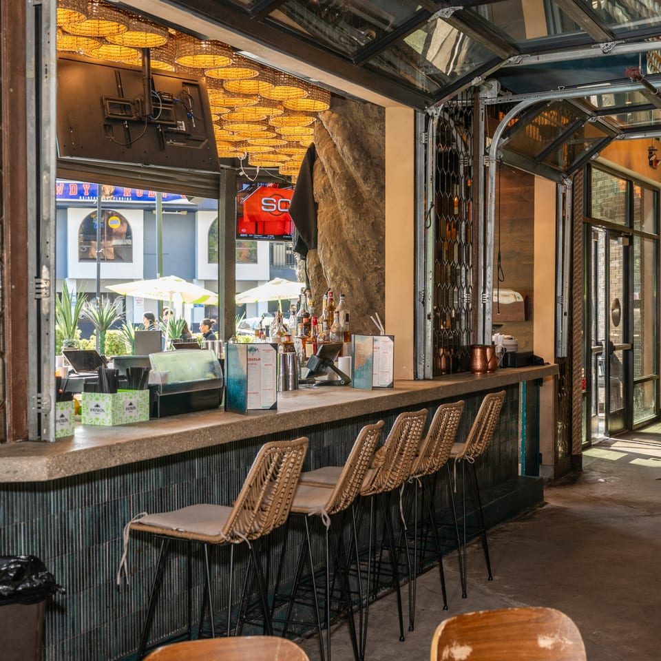 An outdoor bar with wicker stools, bottles, and menus, overlooking a sunny street scene.