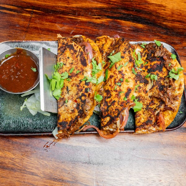 Three golden-brown quesabirria tacos with cilantro, red onions, and a side of dipping sauce on a dark plate.