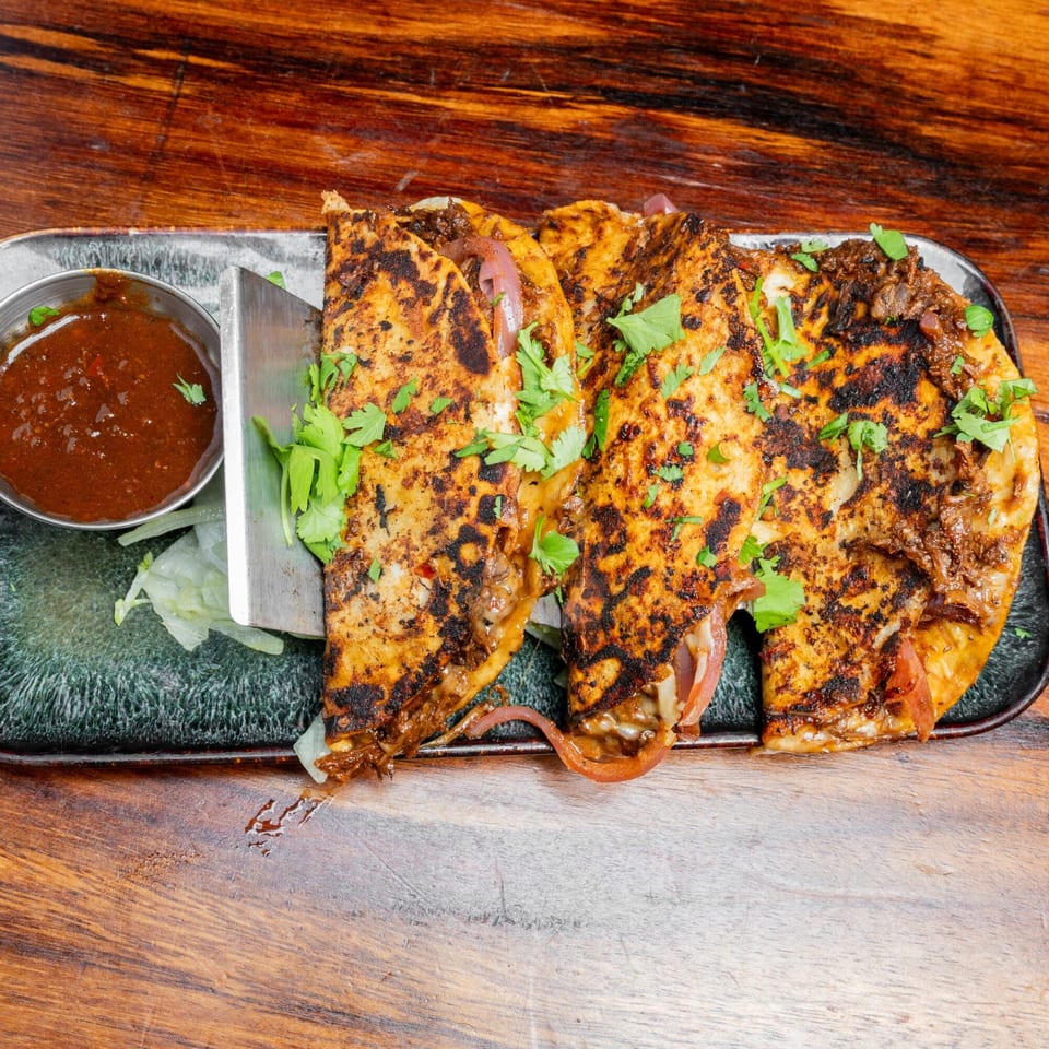 Three golden-brown quesabirria tacos with cilantro, red onions, and a side of dipping sauce on a dark plate.