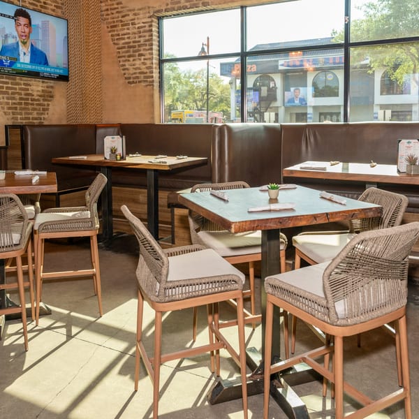 Restaurant interior with high-top tables, woven chairs, brown booths, and a large window overlooking a street.