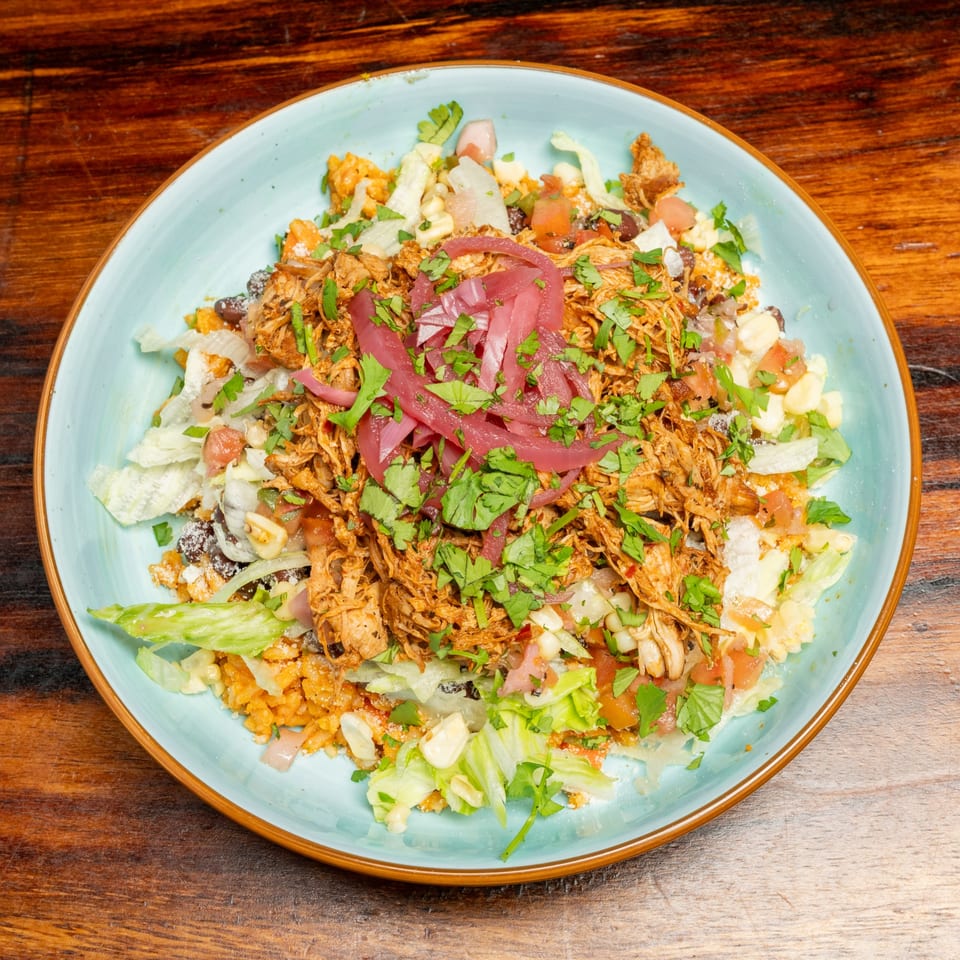 A colorful bowl of shredded chicken, rice, lettuce, corn, and pickled red onions on a wooden table.