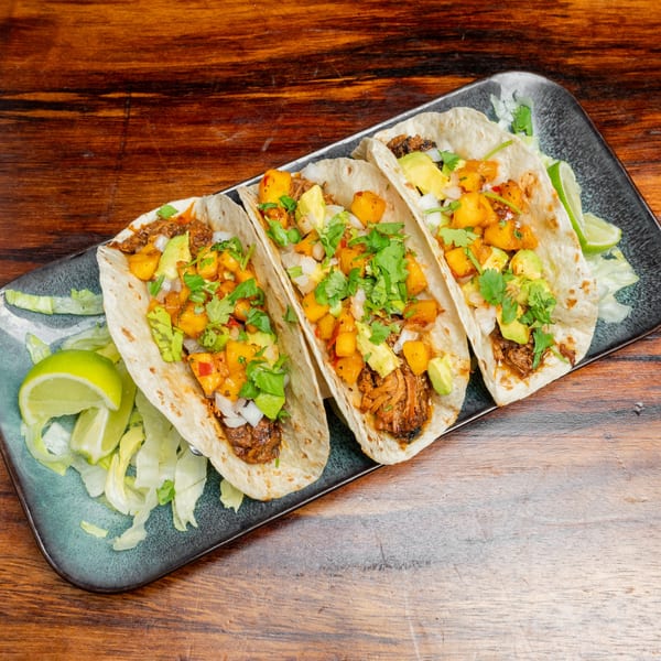 Three tacos with meat, mango salsa, avocado, and cilantro on a dark plate with lime wedges.