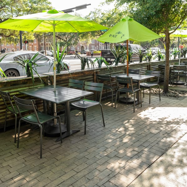 Outdoor patio with green umbrellas, dark tables, and chairs next to a street with cars.