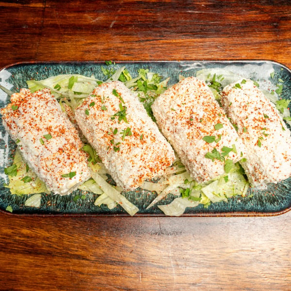 Four elotes, Mexican street corn, on a bed of lettuce, sprinkled with cheese and chili powder.