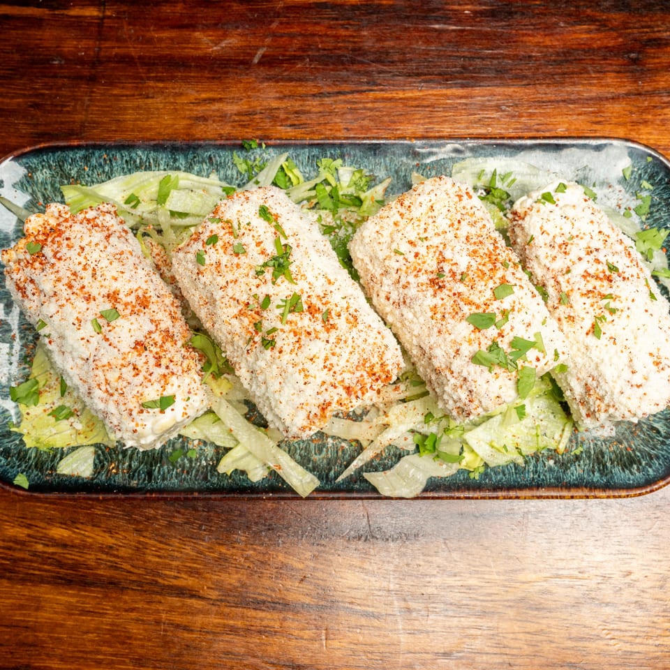 Four elotes, Mexican street corn, on a bed of lettuce, sprinkled with cheese and chili powder.